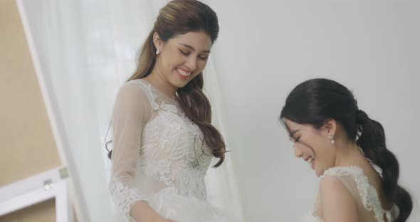 Asian Bridesmaid Helps Bride To Put On The Bride's Wedding Dress.