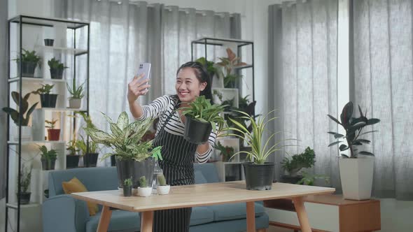 Smiling Asian Woman Holding Plant And Taking Photo By Smartphone At Home