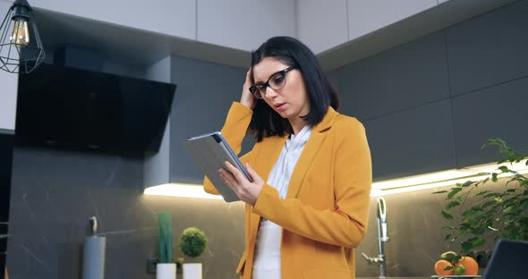 Woman in Glasses and Stylish Orange Jacket Standing in Cozy Kitchen and Using Tablet Device