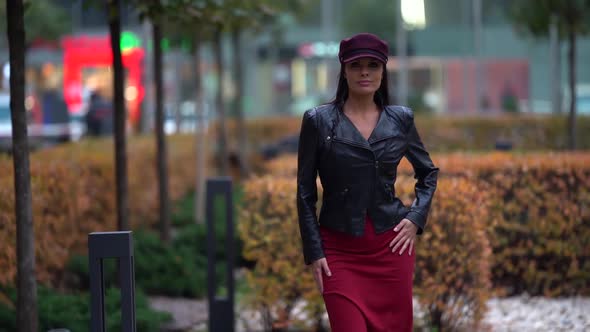 a Brunette in a Black Leather Jacket Red Skirt and a Burgundy Cap Walks Against the Background of a