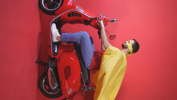 Young Crazy Guy Sitting on a Scooter Moped in a Raincoat and a Superhero Mask Isolated Red