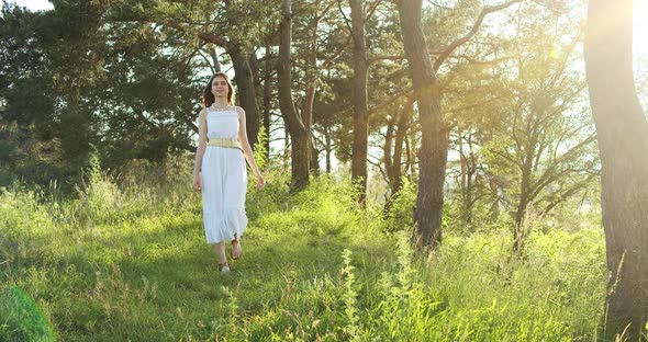 A Cheerful Teenage Girl in Light Dress Walks in a Clearing Between Green Trees in Summer Forest