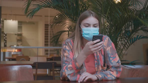 Close Up Portrait of Young Smart Female in a Medical Mask Is Texting on the Phone at Cafe.