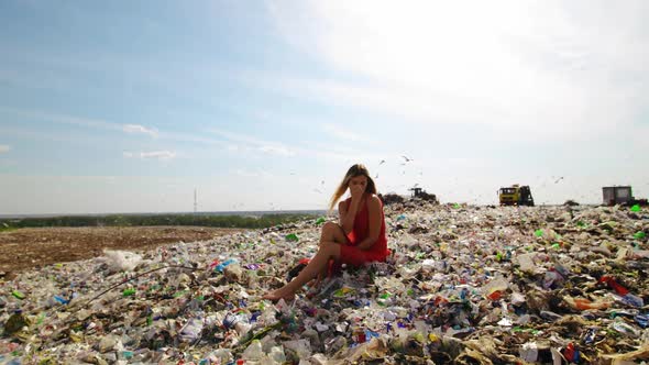Sad Young Woman in Red Dress Sits at Huge Trash Dump with Many Seagulls in Background