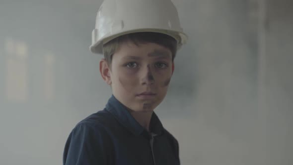 Cute Boy in a Protective Helmet and with Dirty Face Looking at the Camera and Walking in the Smoke