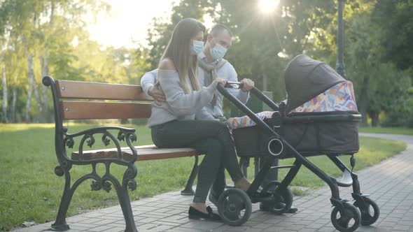 Wide Shot Side View of Couple of Happy Young Parents in Covid-19 Face Masks Sitting in Sunlight in