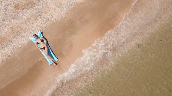 Woman in a Black Swimsuit Lies on a Blue Mattress on a Sandy Beach By the Sea