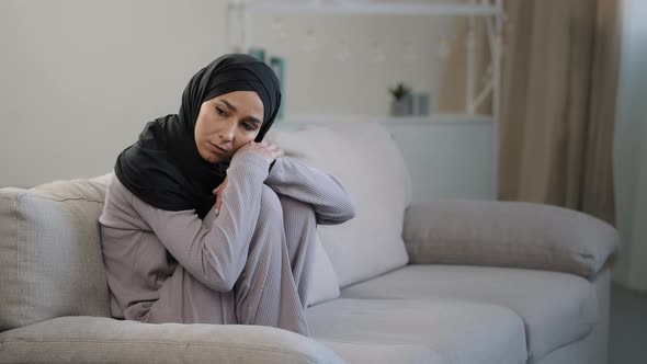 Anxious Sad Young Woman in Black Hijab Sitting on Couch in Living Room Suffering From Psychological