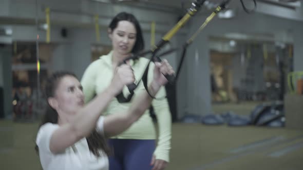 Professional Female Coach Training Young Brunette Woman in Gym Using TRX Straps. Portrait of