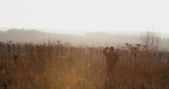 Hunter in Hunting Equipment with Rifle on His Shoulder Walks Through the Field in Sun Light