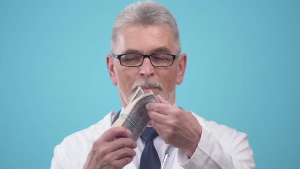 Elderly Doctor Stands on Blue Background and Counts Thick Wad of Dollars and Smiles Front View