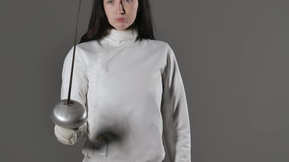 Portrait of a Young Woman Fencer in a White Uniform Posing with a Rapier in Her Hands on a Gray