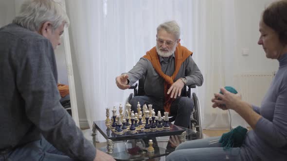 Confident Elderly Man in Eyeglasses Playing Chess with Fellow in Nursing Home As Old Woman Knitting
