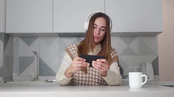 Woman in Headphone Play Game with Smart Phone on Modern Kitchen Background