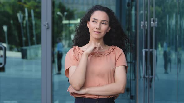 Hispanic Pensive Puzzled Sly Woman Brunette Girl Thinking Stands Outdoors on Background of City