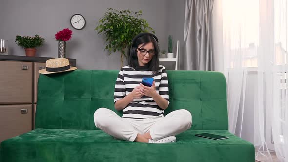 Young Woman in Headphones Sitting on Soft couch in Room and Listening Nice Songs