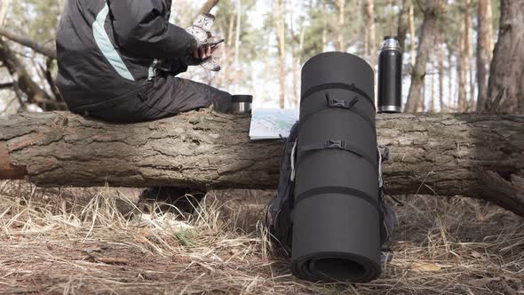 A tourist on a walk in a forest park drinks tea from a thermos and looks at a map on a smartphone.