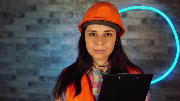 Portrait of Female Construction Worker in Overalls with Clipboard Against Illuminated Wall.