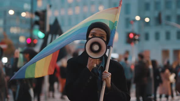 Young Woman with Face Mask Speaking Into the Megaphone While Holding Rainbow Flag in Crowd