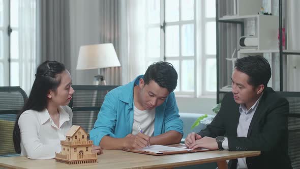 Asian Man With A Woman And Real Estate Agent Signing On House Purchase Contract Paper