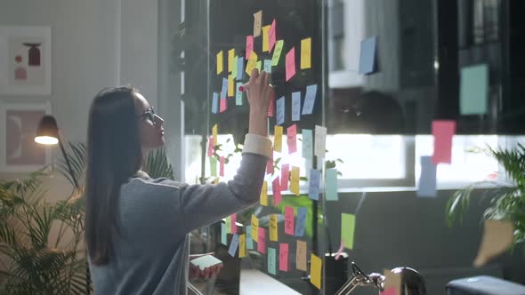 Young Business Woman Brainstorming. Asian Woman Writing Down Ideas On Sticky Notes Attached To Glass