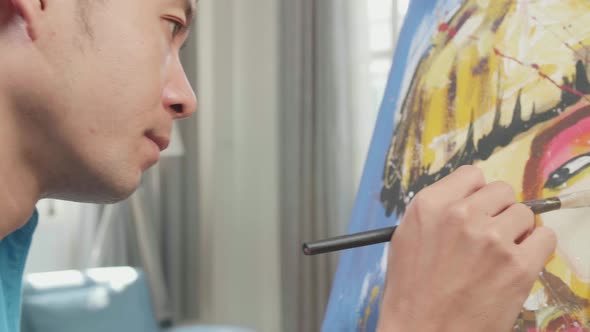 Side View Of An Asian Artist Man Holding Paintbrush Painting A Girl's Face On The Canvas