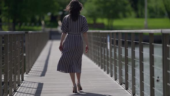 Adult Woman is Walking Alone in Summer Day Strolling Over Bridge in Park Rear View