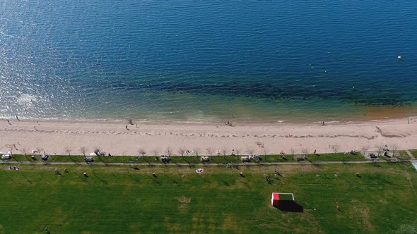 Lake beach with people sunbathing and bathing, garden and walking area. Overhead drone shooting trav