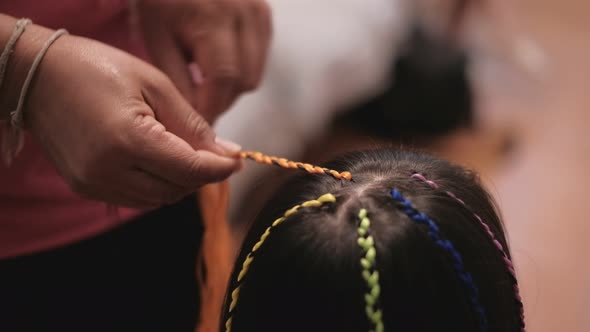 Asian girl is being braided in multi-colored threads by woman's hand.