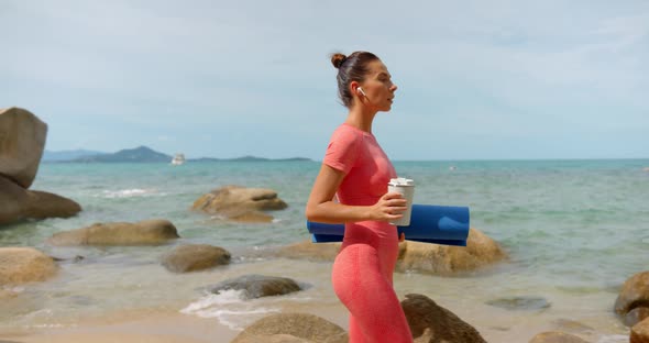 Young Lady in Pink Yoga Suit Walking on Sunny Beach with Bottle of Water and a Yoga Mat in Her Hands