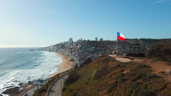 Aerial view passing close to republic of Chile flag flying on Reñaca hilltop revealing downtown city