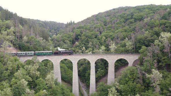 Steam engine train crossing a mountain valley over a stone viaduct.