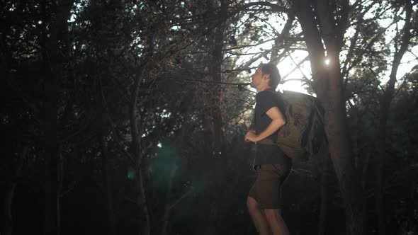 Young Man Hiker with Backpack Standing on Top of Cliff in Summer Mountains in Forest at Sunset and