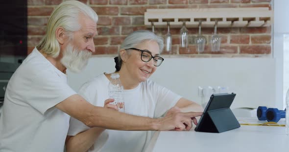 Old bearded Man and His Wife Sitting at Kitchen Table and Watching Interesting Videos