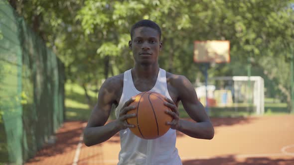 Portrait of Confident African American Sportsman Posing with Ball on Basketball Court. Young Serious