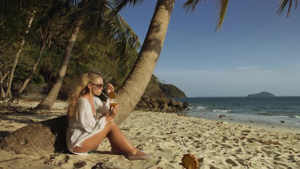 Woman Sitting in White Dress Drinks Pineapple Cocktail Pina Colada on Beach Near Palm