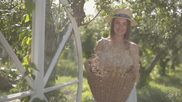 Cute Young Woman in Straw Hat Holding the Wicker Basket with Herbs and Looking at the Camera Smiling