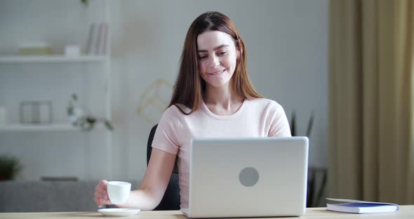 Smiling Happy Young Millennial Woman Sitting at Table at Home Office, Picking Up Hot Cup of Coffee