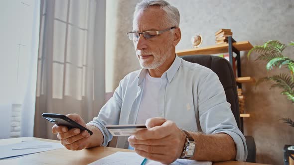 Mature Businessman is Holding Credit Card and Entering Its Number Into Cellphone Shopping Online