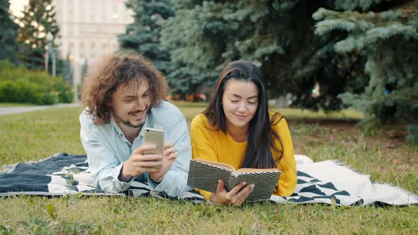 Guy Using Smartphone While Girl Reading Book Relaxing Outdoors in City Park