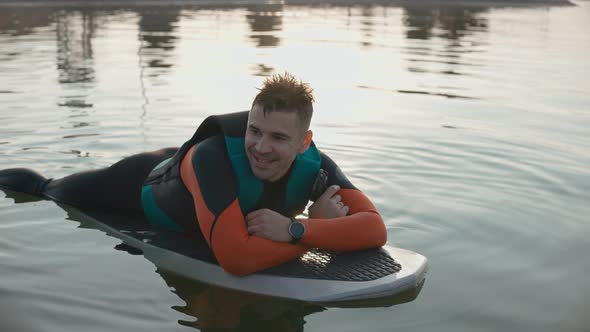 Man Lying and Smiling on a Surfboard Wearing Wetsuit at Warm Golden Sunset