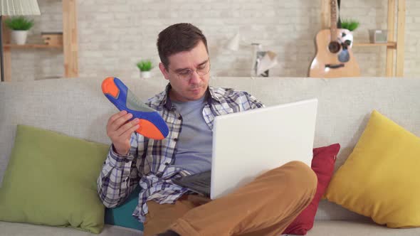 Man Sitting at a Table Uses a Laptop and Holds Orthopedic Insoles in Hand