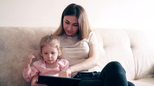 : Young Cute Mother and Little Girl Are Sitting on the Couch and Teaching Using a Tablet Computer