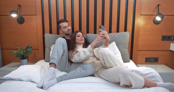 Young Couple Takes a Selfie on a Smartphone Man and a Woman Lying on the Bed and Shoot a Video on a