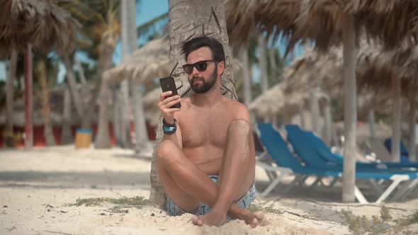 Man Sitting On Palm And Talking On Smartphone On Vacation Lounge Sea. Guy Relaxing On Tropical Beach
