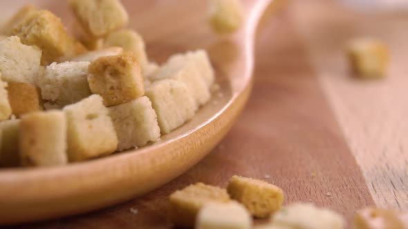 Crispy cubes of toasted wheat bread croutons fall into a wooden spoon and scatter on a wood surface