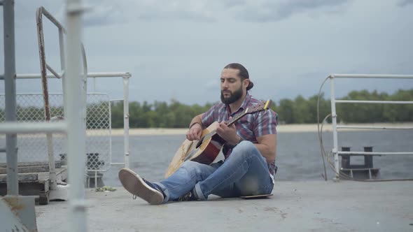 Wide Shot of Thoughtful Caucasian Musician Sitting on Urban Embankment and Playing Guitar. Portrait