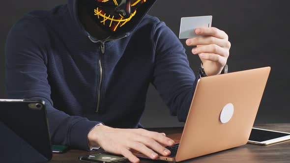 Male Hacker in a Hood Uses Laptop To Access Credit Card in Dark Office Room