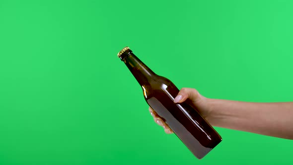A Female Hand Passes a Dark Brown Bottle with Beer or Lemonade to a Male Hand Isolated on a Green