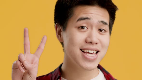 Cheerful Asian Guy Gesturing VSign Smiling Posing Over Yellow Background